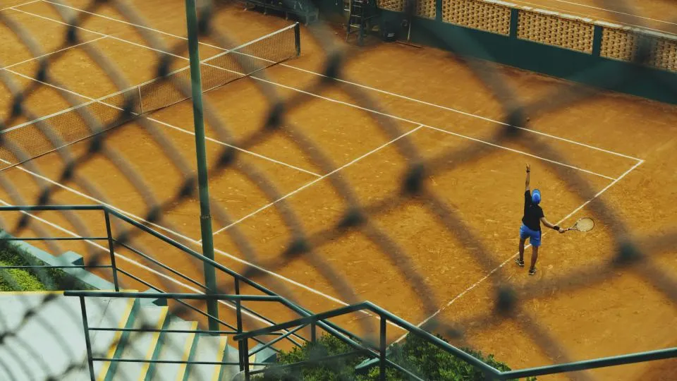 Jugador sirviendo en una pista de tierra batida vista a través de una valla